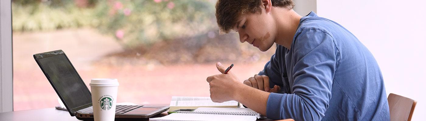 A young male student sits at a high top table working on a laptop in a sunlit room